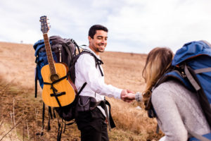 groom helping bride up a mountain. groom has a backpack with a guitar.